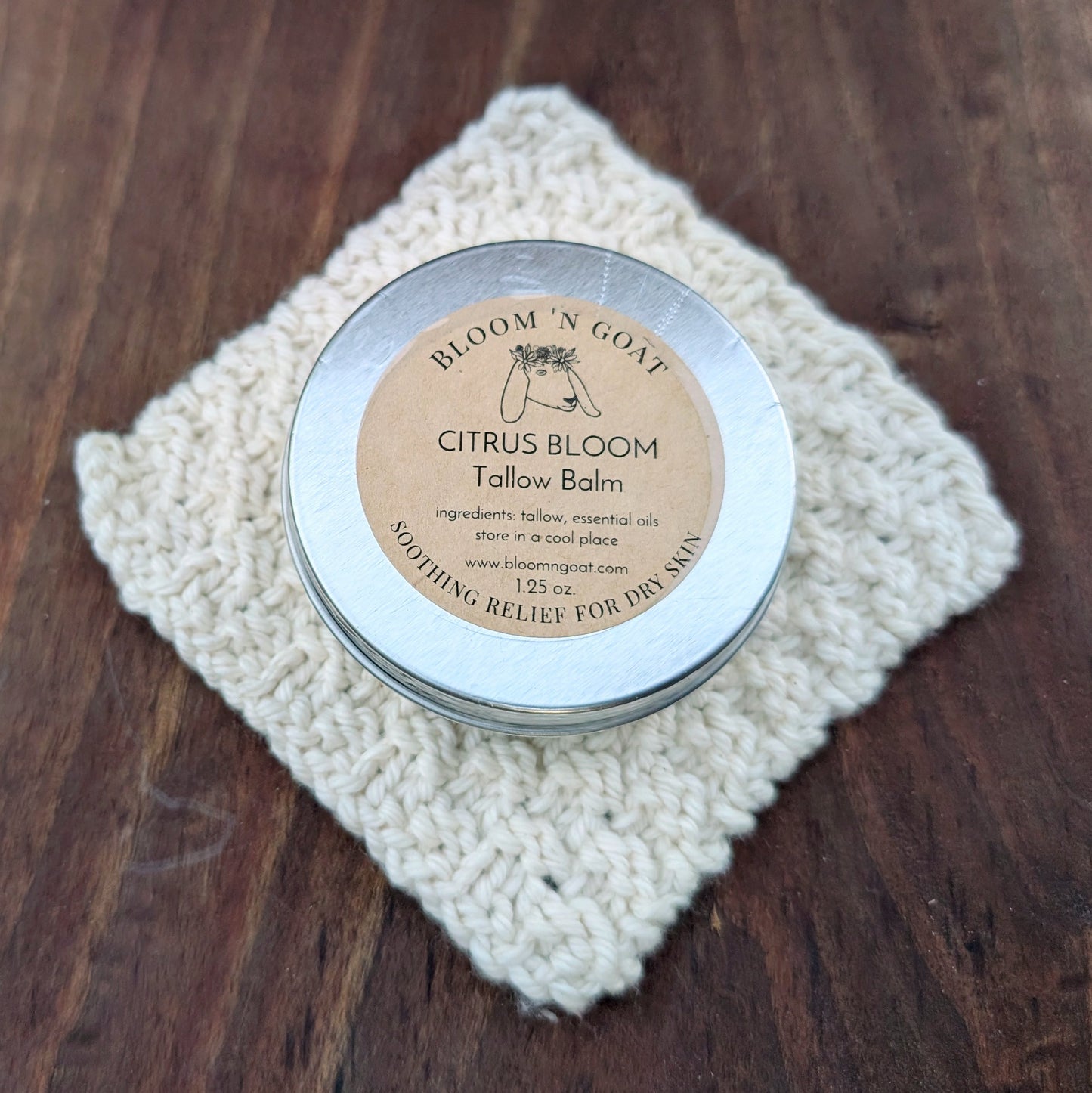 Bloom 'N Goat warm and grounding with frankincense Citrus Bloom Tallow Balm tin on a handknit cloth with a dark walnut wooden background
