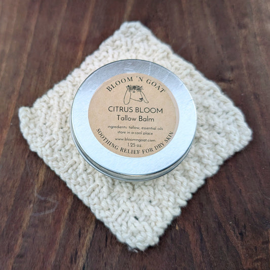 Bloom 'N Goat warm and grounding with frankincense Citrus Bloom Tallow Balm tin on a handknit cloth with a dark walnut wooden background