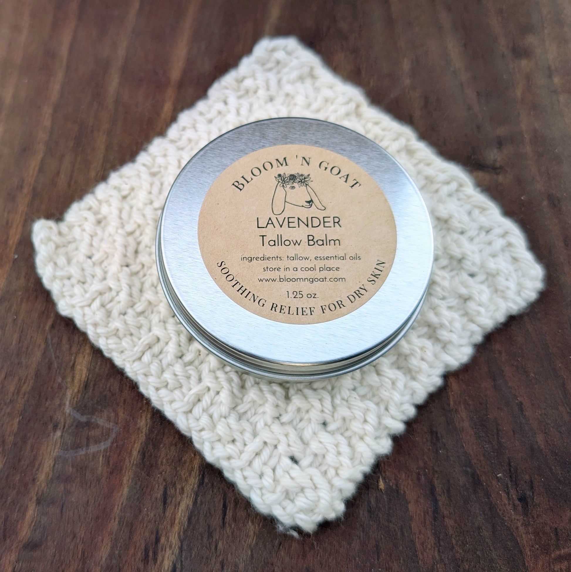 Bloom 'N Goat Lavender calming Tallow Balm tin on a handknit cloth with a dark walnut wooden background