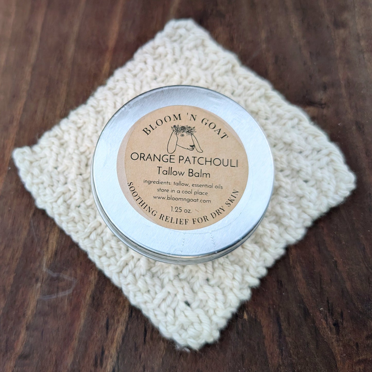 Bloom 'N Goat uplifting and cheerful Orange Patchouli Tallow Balm tin on a handknit cloth with a dark walnut wooden background